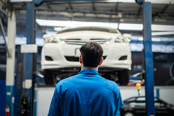 Vehicle service maintenance young handsome man checking car condition on lifter hoist in garage. Automotive mechanic male stand and looking at car seen from behind. Car repair service concept.