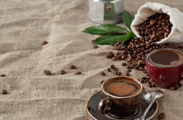 Two colored cups wiht coffee (espresso) and coffee beans in burlap sack on a burlap background. Close up, soft focus, copy space for text. Near coffee leaves and coffee pot.