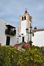 Whitewashed Stone Church And Bell Tower In Betancuria, Fuerteventura With Red Flowers And Green Leaves In Foreground. Blue Sky And Clouds.