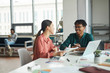 © AnnaStills - Young multiethnic business couple smiling and talkign to each other while sitting at the table with laptop at office