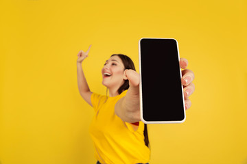 Poster - Showing blank phone's screen. Caucasian woman's portrait isolated on yellow studio background. Beautiful brunette model in casual. Concept of human emotions, facial expression, sales, ad, copyspace.