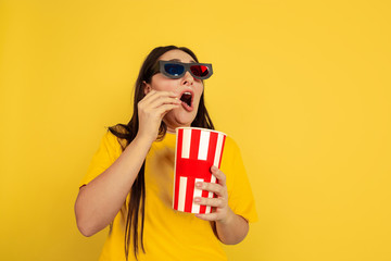 Wall Mural - Watching cinema in 3D-eyewear with popcorn. Caucasian woman on yellow studio background. Beautiful brunette model in casual style. Concept of human emotions, facial expression, sales, ad, copyspace.