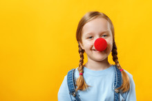 Happy Funny Child Clown With A Red Nose. Cheerful Little Girl On A Yellow Background. April 1st Fool's Day. Copy Space