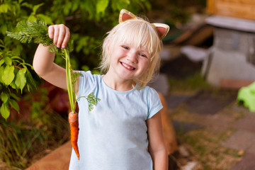 Pretty happy blond toddler girl wearing cat's headband picking fresh carrot, gardening outdoors on a sunny summer day