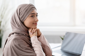 Wall Mural - Smiling muslim businesswoman wearing headscarf, sitting at workplace and looking away