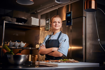 Confident and slightly smiling female chef standing in a dark kitchen next to cutting board with vegetables on it, wearing apron and denim shirt, posing for the camera, reality show look