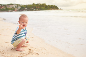 Wall Mural - Cute little Asian toddler baby boy child on beach with dirty hands covered with wet sand. Family travel, water outdoor activity on summer beach vacation with children, Sensory play with sand concept