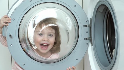 close up of little funny 3 year old girl plays with hatch of washing machine. child looks through ha