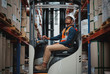Happy forklift driver focused on carefully transporting stock from shelves around the floor of a large warehouse wearing a white helmet and vest