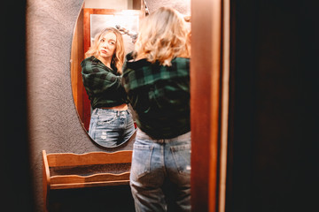 Wall Mural - Young woman using curling iron looking at herself in the mirror at home preparing to go out