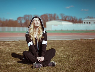 Wall Mural - Young woman in a dark brown sports overalls with a hood sits on grass