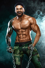 Wall Mural - Athlete bodybuilder on black background with lights and smoke. Men fashion. Portrait of a brutal bearded man topless with chains.