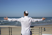 Young Jew In Kippah Hat Stands With Outstretched Arms On A Background Of Blue Sea And Blue Sky