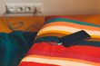 Black smart phone is charged on the pillow in the bed with colored linen.
