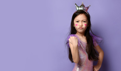 Asian Korean kid girl princess in party dress and crown and with a painted red hearts sign on cheeks makes force gesture