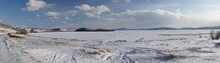 Car Tracks Lead From The Shore To A Snowy Frozen Large Lake With Forest Mountains On The Horizon In The Winter Sunny Day.