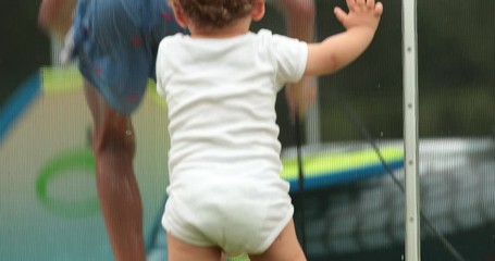 Wall Mural - Infant baby leaning on pool fence looking at children play inside swimming pool water during summer day