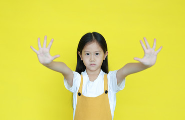 Asian little child girl doing stop sing with two palm of hands. Child show warning expression isolated on yellow background. Negative and serious gesture concept. Selective focus at face