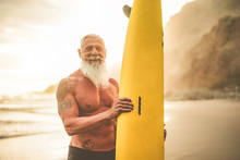 Tattooed Senior Surfer Holding Surf Board On The Beach At Sunset - Happy Old Guy Having Fun Doing Extreme Sport - Joyful Elderly Concept - Focus On His Face