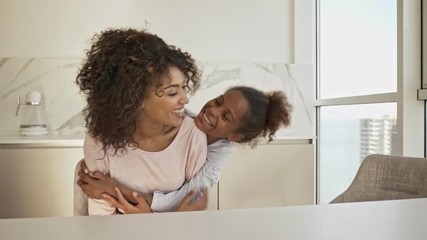 Wall Mural - Pleased little girl hugging behind her mother which sitting by the table on kitchen at home