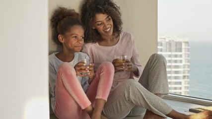 Wall Mural - Joyful african woman and her satisfied little pretty daughter drinking juice while sitting on the floor near window at home