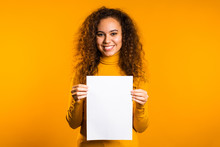 Pretty Curly Woman Holding White Vertical A4 Paper Poster. Copy Space. Smiling Trendy Girl On Yellow Background.