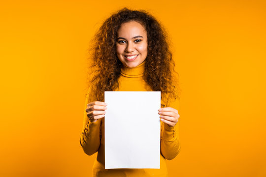 Pretty curly woman holding white vertical a4 paper poster. Copy space. Smiling trendy girl on yellow background.