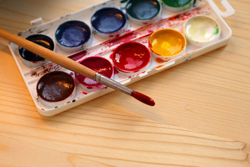 Wet brush for drawing with iron tip and watercolors for children are left on wood brown table. Single brush lies on top of palette of colorful paints for painting. Closeup. Selective focus.