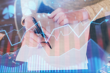 Hand taking notes in notepad. Forex chart holograms in front. Concept of research. Multi exposure