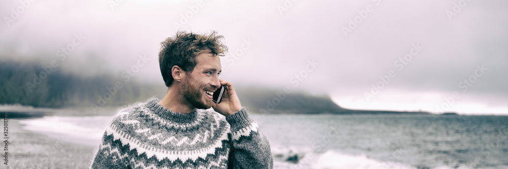 Young man walking on beach talking on the mobile phone having conversation speaking to someone during nature winter walk panoramic banner outdoor lifestyle - obrazy, fototapety, plakaty 