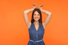 I'm Bunny! Smiling Funny Amusing Brunette Woman In Denim Dress Showing Rabbit Ears Gesture On Head And Looking With Childish Carefree Expression. Indoor Studio Shot Isolated On Orange Background
