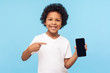 © khosrork - Mobile app for children. Happy excited cheerful little boy with curly hair in T-shirt pointing at cell phone and smiling at camera, showing telephone. indoor studio shot isolated on blue background