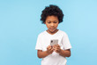© khosrork - Serious attentive cute little boy with curly hair in T-shirt reading typing message on smartphone, browsing on children mobile application using cellphone. studio shot isolated on blue background