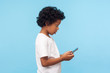 Side view of astonished nice preschool boy with curly hair reading message on smartphone and expressing amazement shock, surprised by mobile application, using cellphone. studio shot, blue background