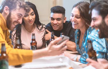 Group of smiling young people watching video on smartphone and drinking beers in a house. Multiracial friends having fun using smartphone, social network apps and new technologies concept.