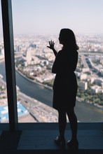 Silhouette Of Young Cute Girl Who Pressed Against The Glass And Admiring The Panoramic View Of City From The Tall Building. Selective Focus On Hand. Blurred Background.