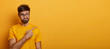 © WHstudio Leushin N - Doubtful man points at upper right corner, feels hesitant about something, purses lips with confusement, poses against yellow backgroud, wears casual clothes. Advertising and people concept.
