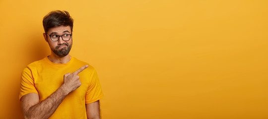 Doubtful man points at upper right corner, feels hesitant about something, purses lips with confusement, poses against yellow backgroud, wears casual clothes. Advertising and people concept.