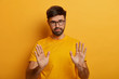 © WHstudio Leushin N - Slow down. Serious looking displeased bearded man shows stop gesture, asks to hold horses, keeps palms towards camera, says take it easy and control your behaviour, stands against yellow background