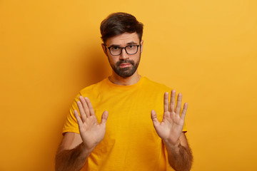 Slow down. Serious looking displeased bearded man shows stop gesture, asks to hold horses, keeps palms towards camera, says take it easy and control your behaviour, stands against yellow background