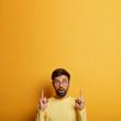 Photo of astonished unshaven man makes annoncement, points index fingers above, demonstrates blank space, good sale offer, recommends service, dressed in casual clothes, poses over yellow wall