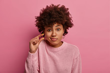 Headshot Of Curly Haired Woman Discusses Something Very Small, Shapes Something Very Tiny, Purses Lips, Dressed In Casual Jumper, Isolated On Pink Background, Asks Tiny Object, Makes Small Gesture