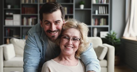 Wall Mural - Smiling 30s son looking at camera hugging older mom indoors