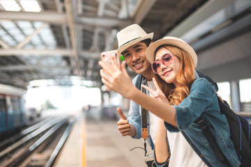 Happy Asian couple traveler holding a mobile phone in station and waiting for train in vacation time.Two Asian Tourists With Backpacks Train travel in Sightseeing City Thailand.