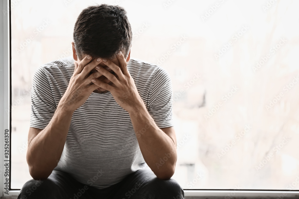 Depressed young man near window - obrazy, fototapety, plakaty 