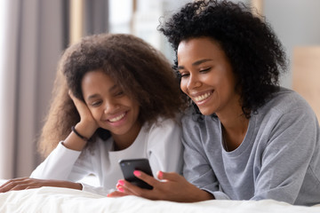 Wall Mural - Happy african mother and daughter using smartphone lying on bed