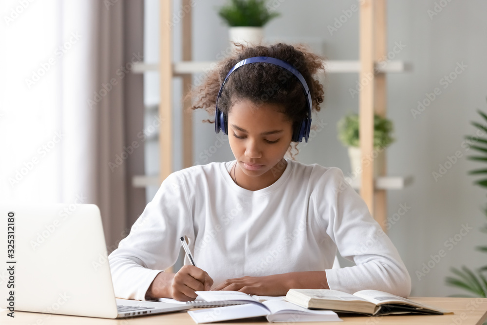 Focused african african teen girl wearing headphones writing notes studying - obrazy, fototapety, plakaty 