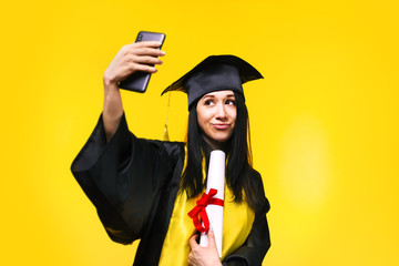 graduate woman makes selfie photo on phone over yellow background, concept of successful completion of studies,