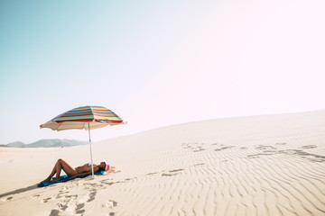 Wall Mural - Beautiful desert dunes beach and caucasian female lay down enjoying a sunbath under a coloured umbrella - concept of summer holiday vacation ant travel tourist under the sun with mountains background