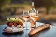 Bottle of rose wine and two full glasses of wine on table in heart of Provence, France with french bread, cheese, ham, grapes and peaches with olive trees on background in sunset. Travel in Provence.